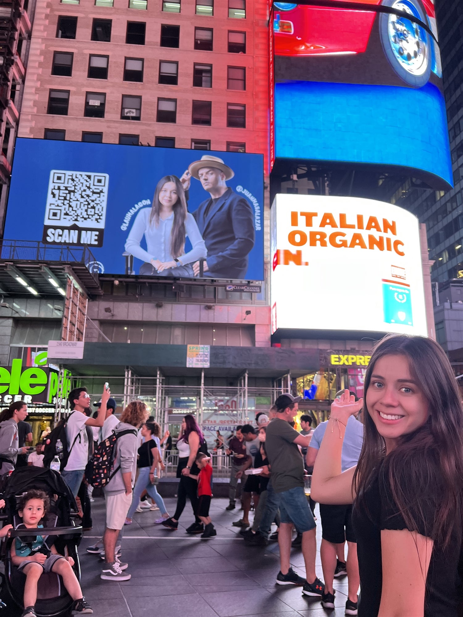 A woman smiles and points at a digital billboard displaying her own image in a busy urban area with people and electronic signs.