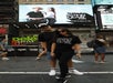 A man and woman in black outfits pose together in Times Square, New York, with a matching "Welcome to Thistimesquare.com" shirt and billboard in the background.