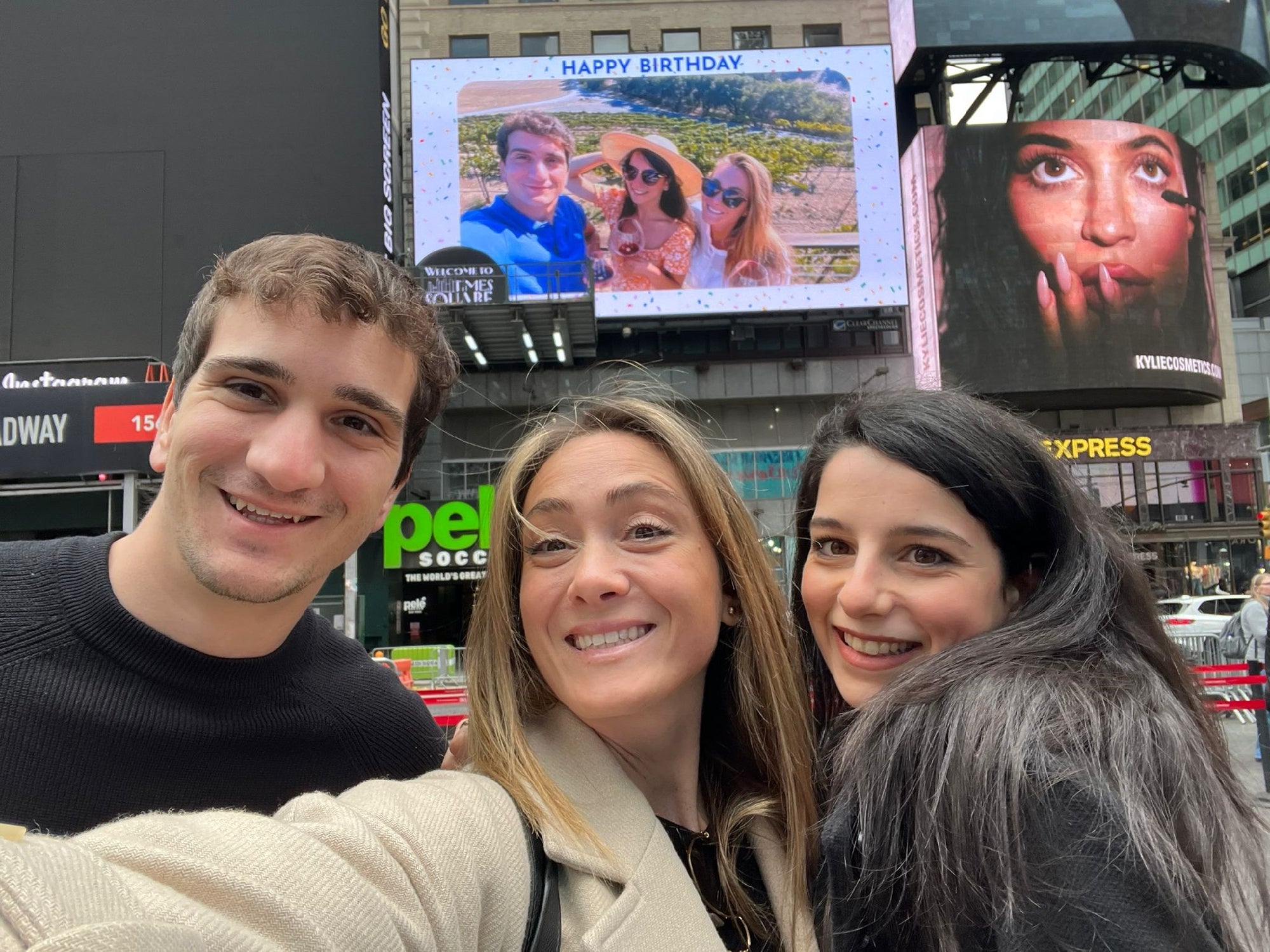 Three people smiling for a selfie in Times Square, with a large “Happy Birthday” billboard and another advertisement visible in the background.