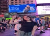 A young woman smiles and points upward in a busy Times Square scene, with a large digital billboard and crowds of people in the background.