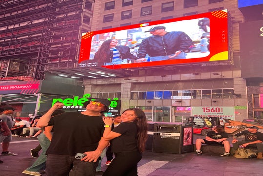 Two people pose and smile in Times Square at night, with their image displayed on a large digital billboard behind them. Other people and bright signs are visible in the background.