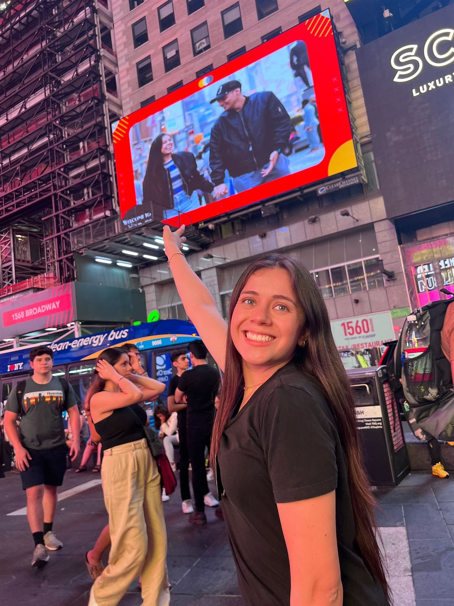 A woman in Times Square smiles and points up at a large screen displaying a photo of two people. Several pedestrians and buildings are visible in the background.