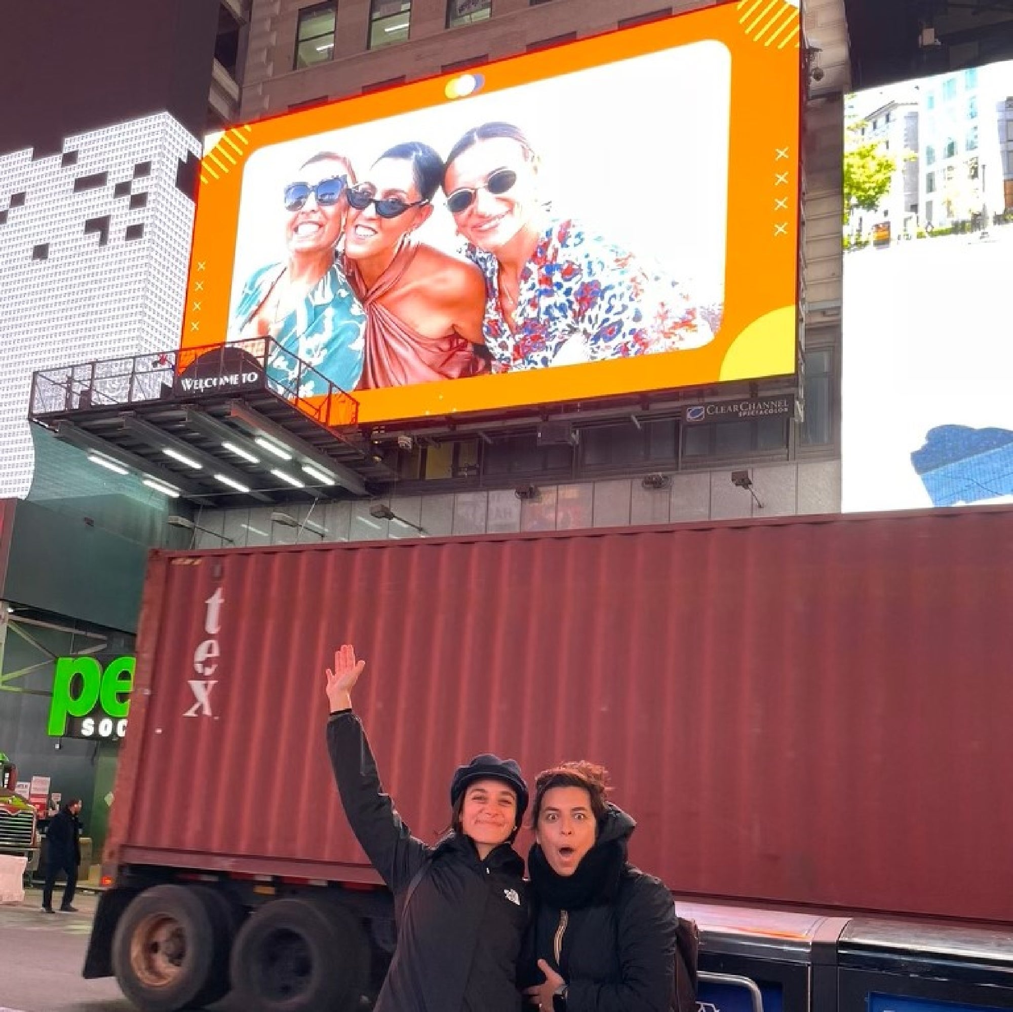 Two people stand on a city street pointing excitedly at a large electronic billboard above them displaying a photo of three smiling people.