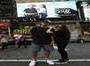 Two people pose in front of a digital billboard in Times Square, New York City. The billboard displays an image of a man and woman standing together.