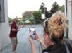 A woman takes a photo of a man posing and smiling outdoors; the man stands in the background while she holds up a smartphone in the foreground.