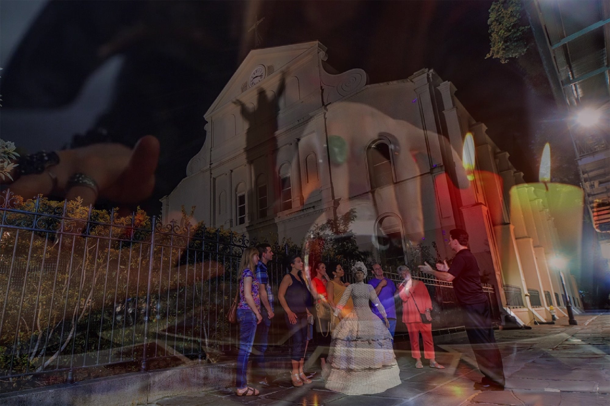 A group of people stand outside a historic building at night, while a person's hand and lit candles are superimposed over the scene.
