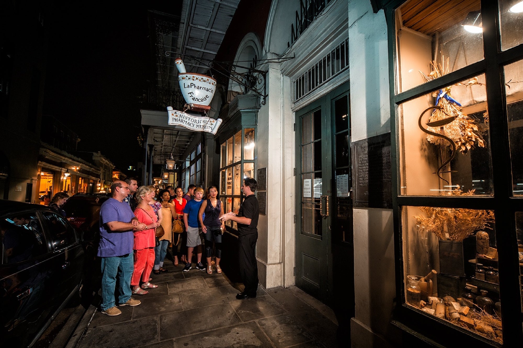 A group of people stands on a sidewalk at night, listening to a guide outside a lit storefront in an urban area.