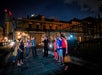 A group of people stand on a sidewalk at night, listening to a guide speak in front of a historic building with iron fencing.