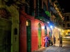 A group of people stand and talk on a sidewalk at night outside colorful, old-style buildings with balconies and green doors under bright streetlights.