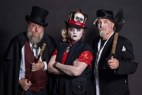 Three people in Victorian Gothic attire pose against a dark backdrop. The person in the center wears a hat with a skull, flanked by two others in top hats and holding a cane and shovel.
