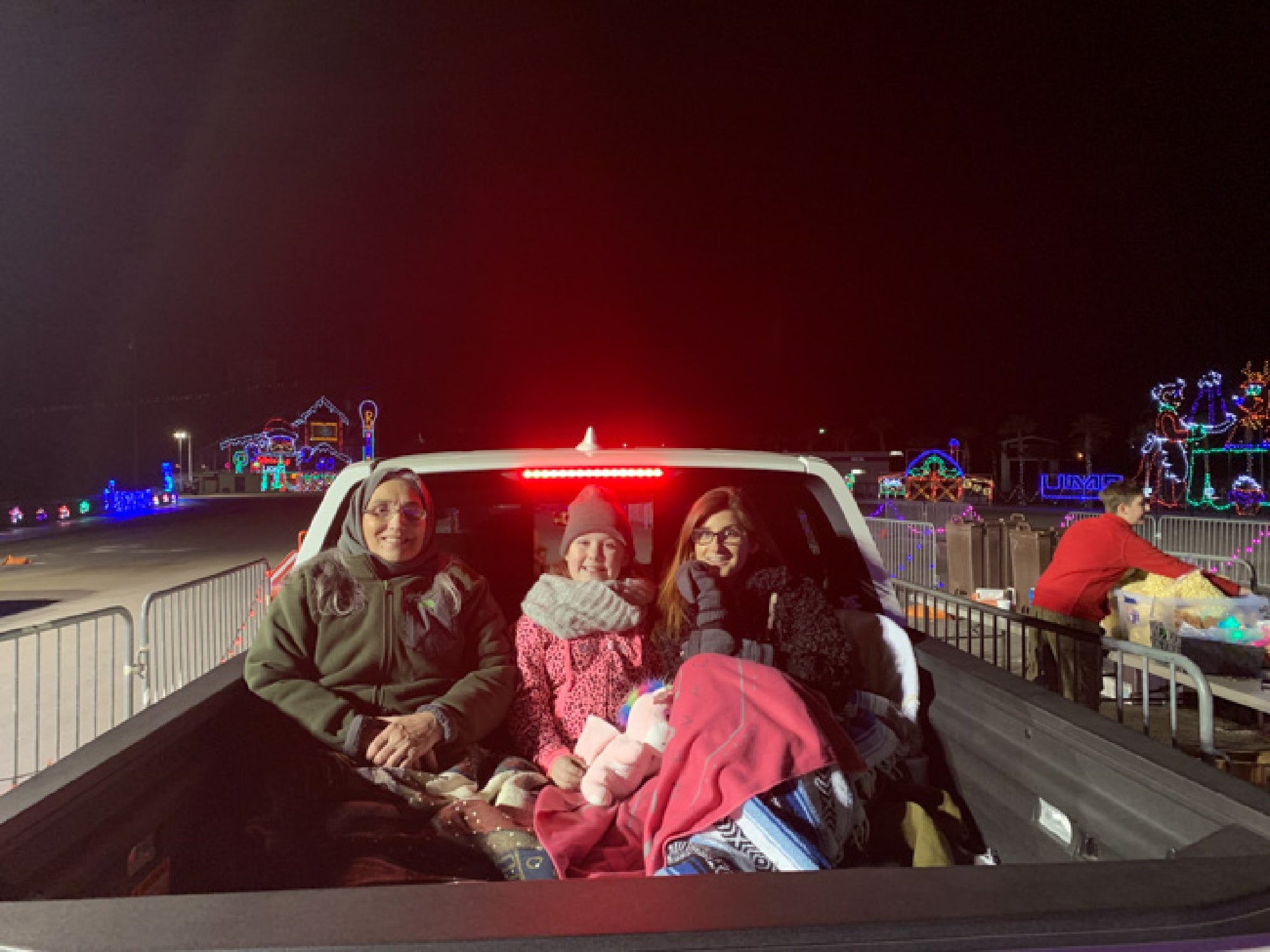 Three people sit bundled up in the back of a truck at night, surrounded by colorful holiday lights.