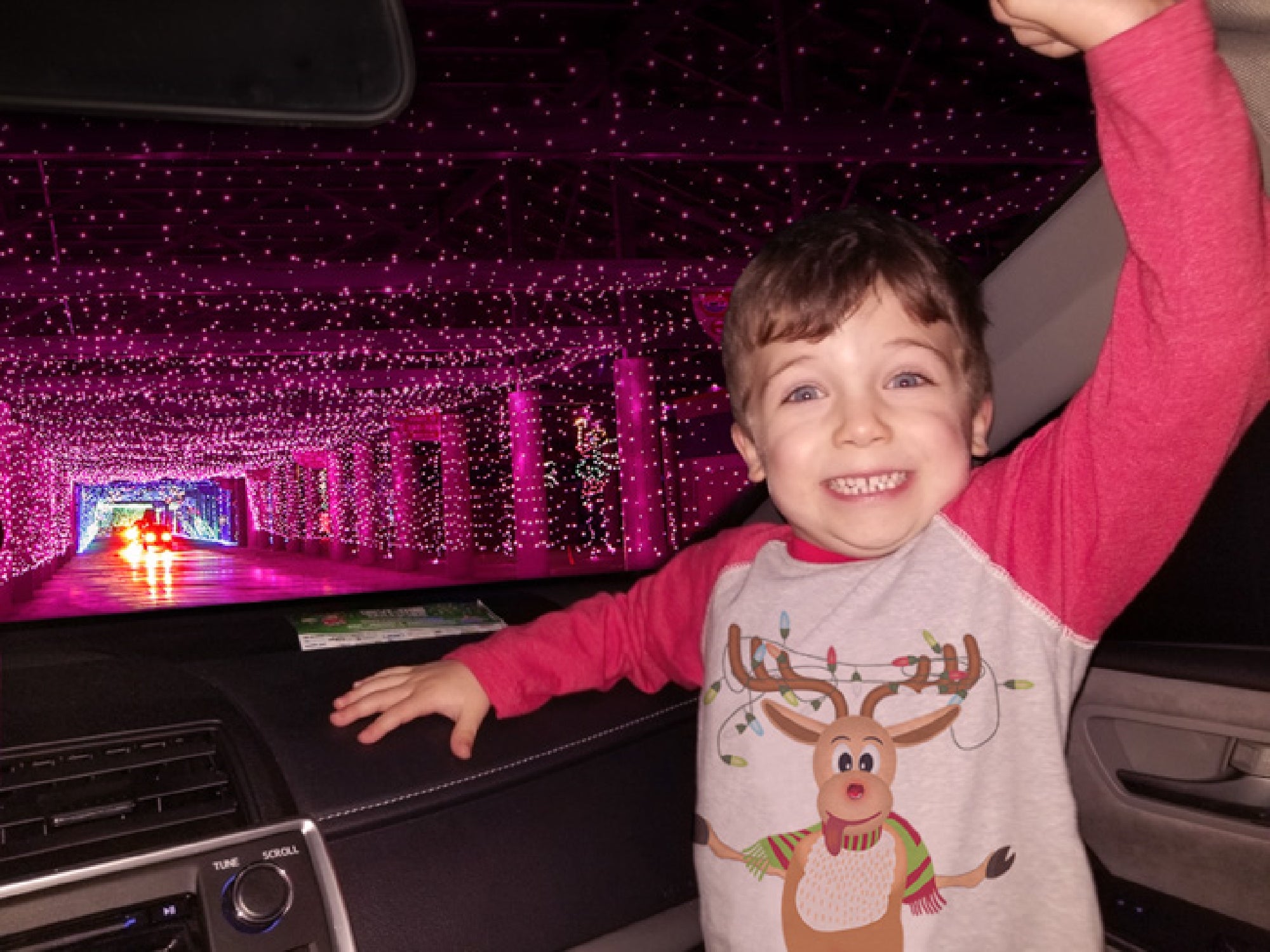 Child smiling inside a car, surrounded by pink holiday lights, wearing a red and gray reindeer shirt.