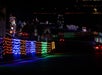 Colorful holiday lights illuminate a dark street with festive displays, including music notes and a radio station sign, creating a vibrant nighttime scene.