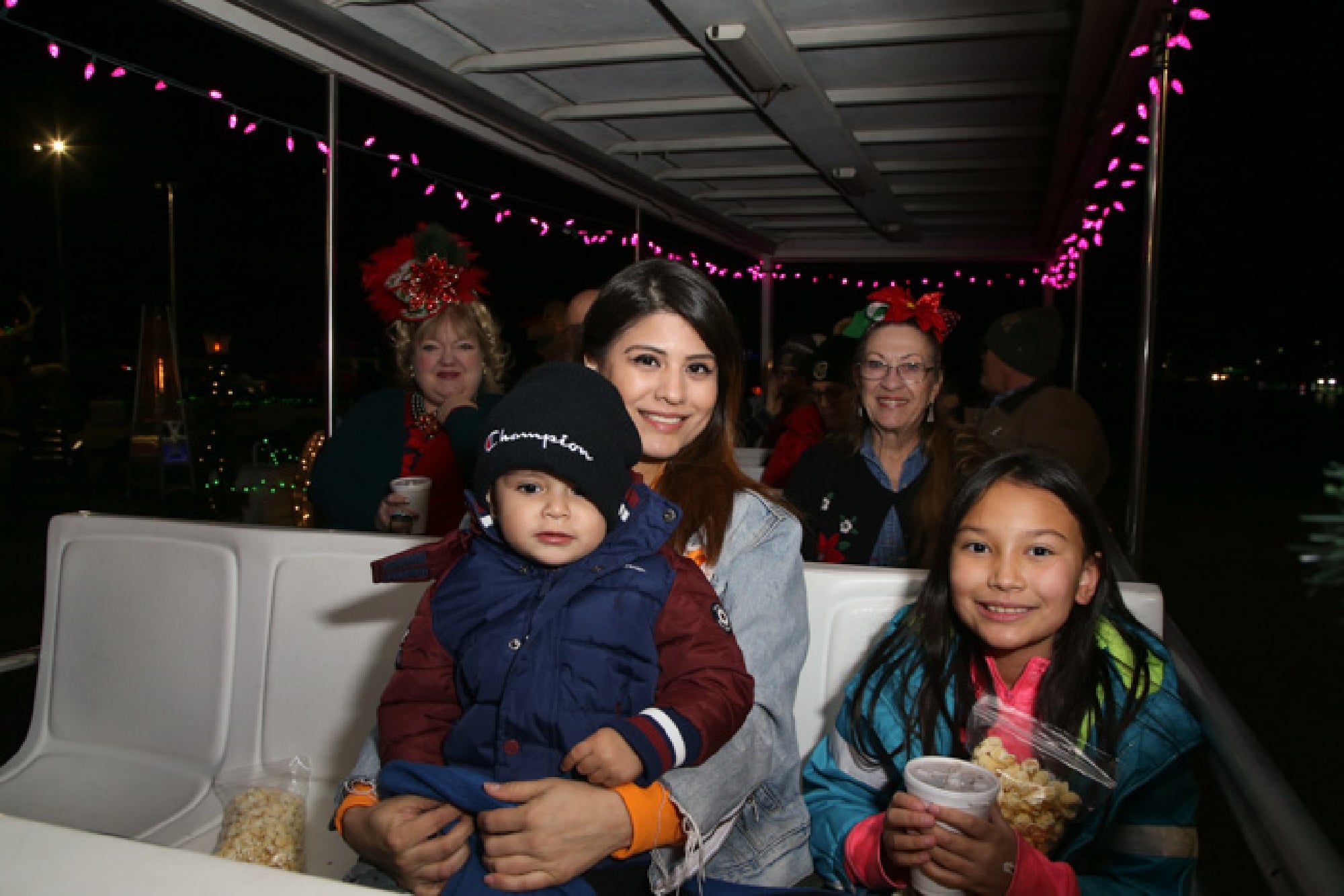 A group of people, including children and adults, sit together on a lit-up ride at night, some holding snacks.