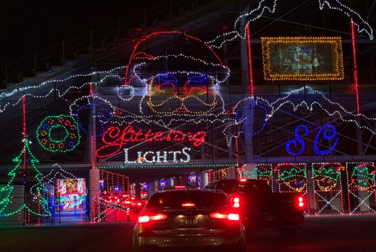 Cars driving through a holiday light display featuring a large Santa face, colorful decorations, and the words "Festival of Lights" illuminated in various colors.