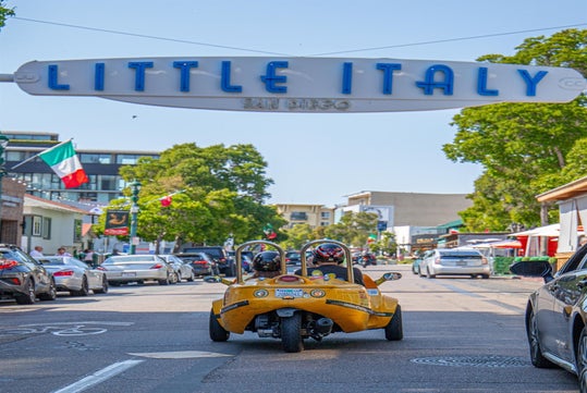 A yellow three-wheeled vehicle drives under a "Little Italy San Diego" sign, with trees and buildings in the background.