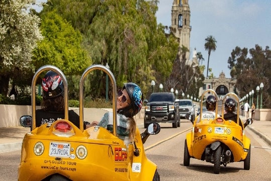 Two yellow GoCars with passengers wearing helmets driving them down a busy road in San Diego with palm trees to their right.
