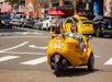 View of a yellow GoCar from behind on the street with two tourists wearing helmets driving it and a line of cars in front of it.