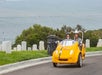 A man and woman wearing helmets and driving a yellow GoCar next to a cemetery with water in the background.