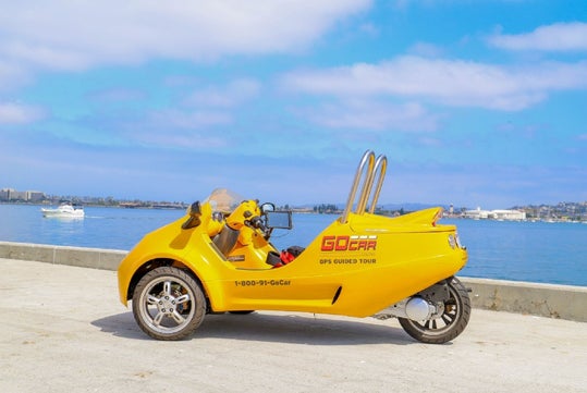 A yellow three-wheeled GoCar vehicle is parked beside a waterfront on a sunny day, with water and distant buildings visible in the background.