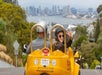 View of the back of a GoCar with a man in a silver helmet driving and a woman in a black helmet turned back with her hand up showing a peace sign.