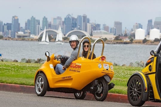 Tourists wearing helmets sitting in a yellow GoCar parked on the side of the road with boat in the water and the city in the background.