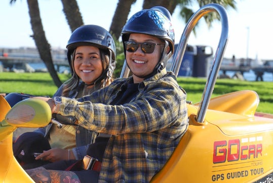 Two people wearing helmets sit in a yellow GoCar for a GPS guided tour, smiling at the camera with palm trees and grass in the background.