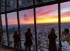 Several people stand by large windows overlooking a cityscape at sunset, with a “West” sign indicating direction on the glass.