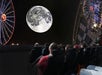 People seated in a planetarium watch a projection of the moon with an illustration of a rabbit drawn on its surface; Ferris wheels are visible on either side.