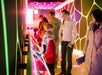 A group of adults and children observe interactive displays in a brightly lit, modern exhibit space with colorful, geometric wall patterns.