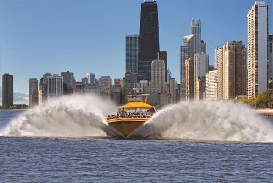 A yellow speedboat creates large waves as it moves across the water with a city skyline of tall buildings in the background.