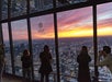 People stand by large windows in a high-rise observation deck, photographing a colorful sunset over a cityscape labeled "WEST.