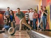 A group of people stand and sit by a bowling lane, watching as a man in the foreground bowls a ball down the lane.