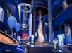 A science museum exhibit features a large vortex tornado display, blue hot air balloons, interactive stations, and colorful lighting in a modern, spacious room.
