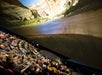 A large group of people seated in a theater watch a curved, panoramic screen displaying a scenic river flowing between rocky cliffs.