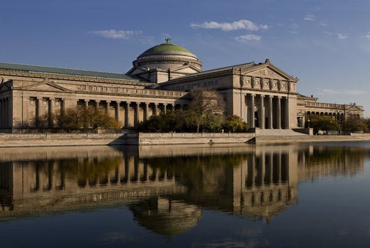 Neoclassical building with columns and a large dome, reflected in a calm body of water under a clear blue sky.