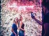 Two women pose in front of a white flower wall with a neon Madame Tussauds sign while a person takes their photo with a smartphone.