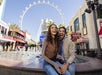 Two people sit and smile in front of a fountain at an outdoor shopping area, with a large Ferris wheel and various storefronts in the background.
