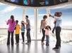 People stand in an observation deck looking out large windows at a cityscape; some are pointing and others are interacting with children.