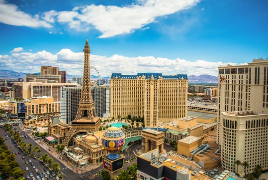 A daytime view of the Las Vegas Strip featuring hotel towers, the replica Eiffel Tower, and nearby buildings under a blue sky with clouds.