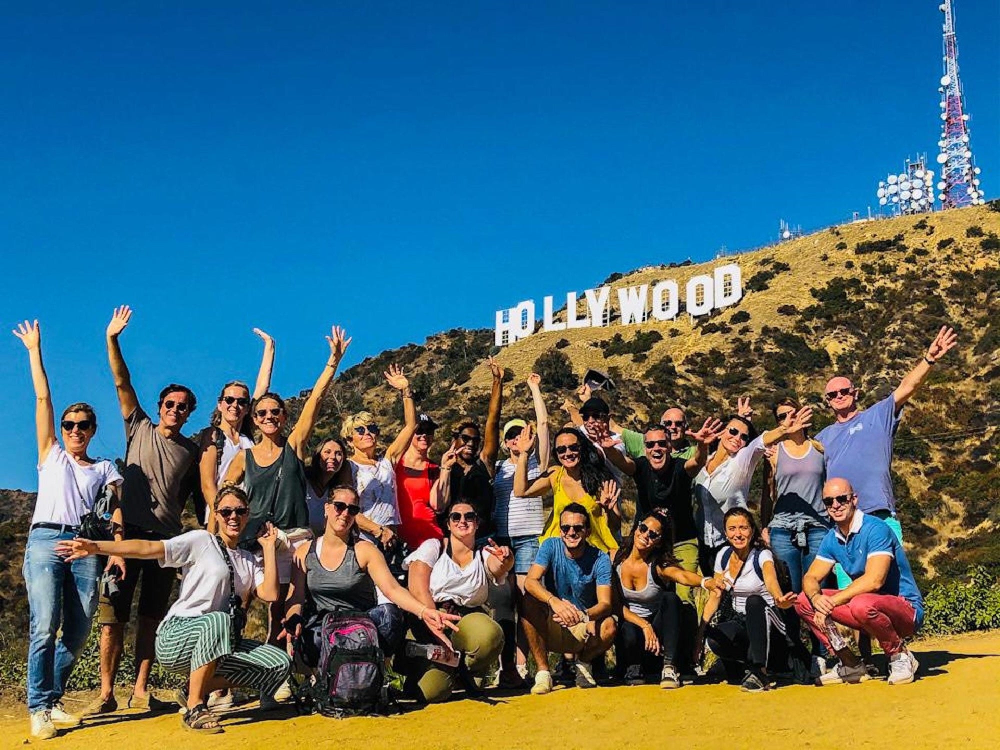 A group of people posing enthusiastically in front of the Hollywood sign on a sunny day, with clear blue skies and a hill in the background.