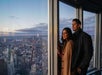 A man and woman stand by a large window in a tall building, overlooking a cityscape at sunset.