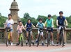 Six people, including four children and two adults, stand with bicycles wearing helmets on a bridge with decorative stonework and trees in the background.