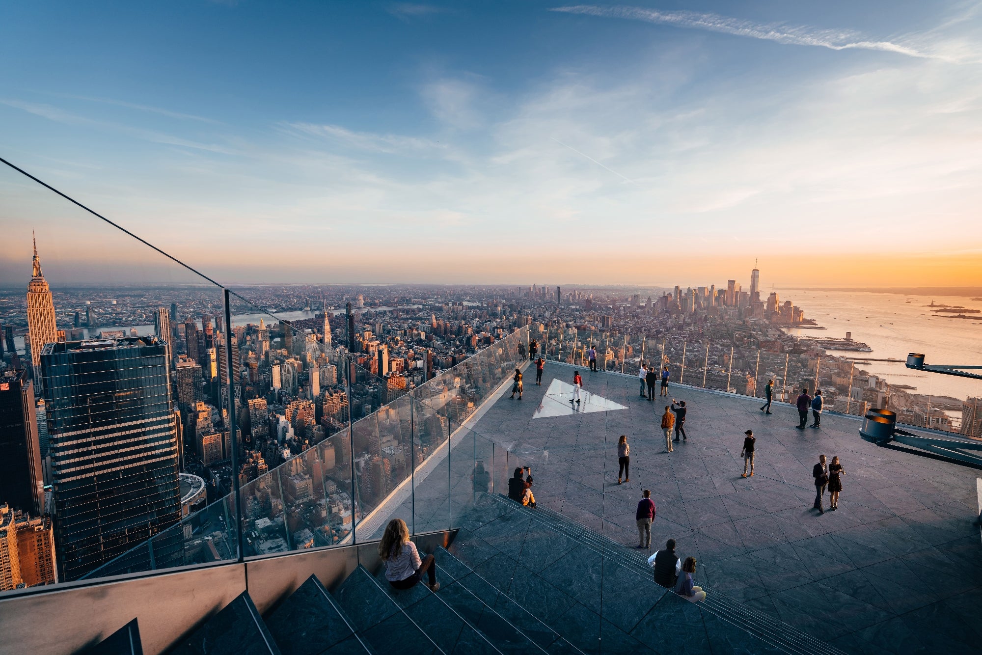 People stand on an outdoor observation deck with glass railings, overlooking the New York City skyline at sunset.