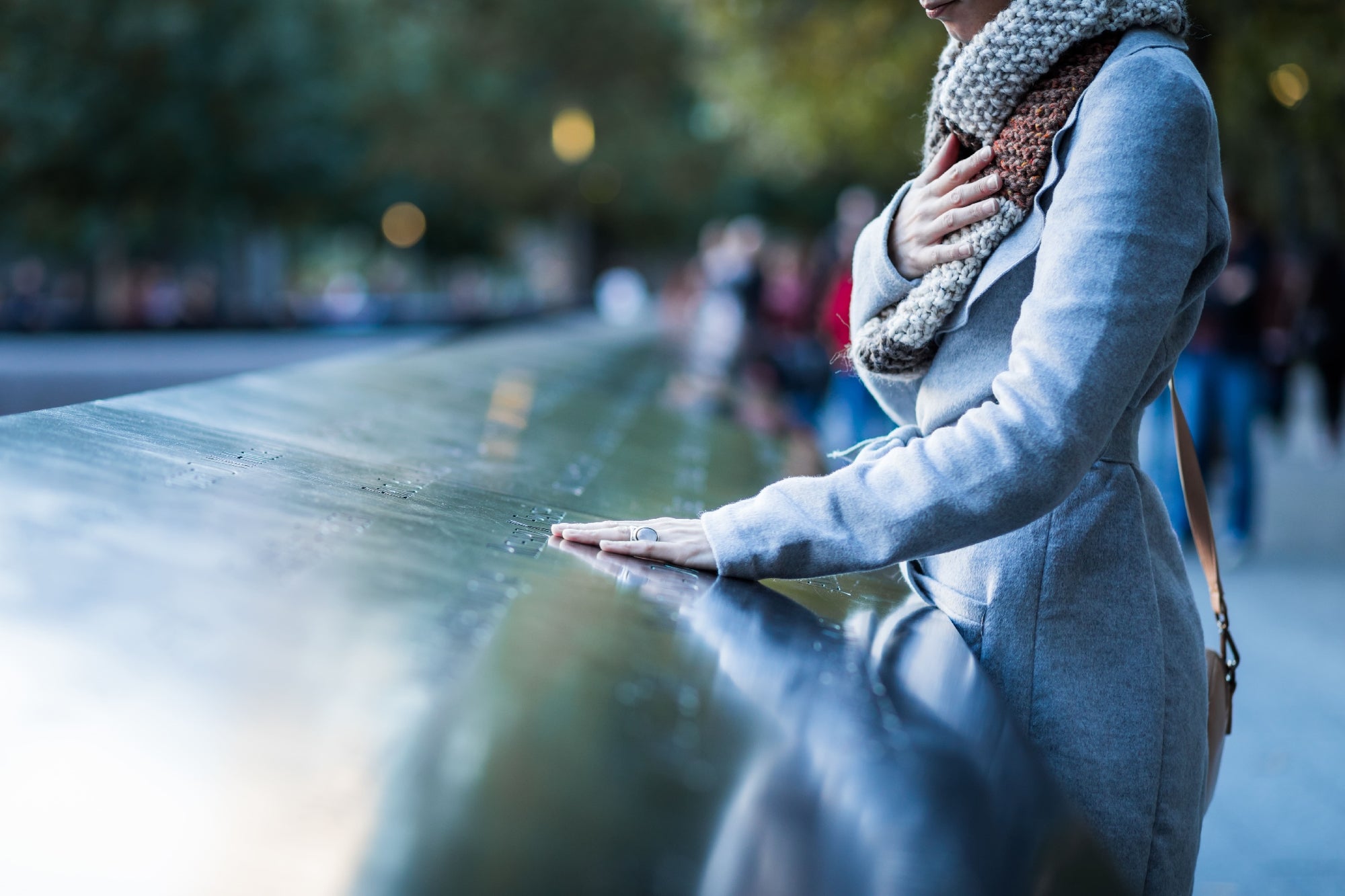 Person wearing a scarf and coat stands with one hand on a memorial wall and the other on their chest, appearing reflective. Trees and blurred people are visible in the background.