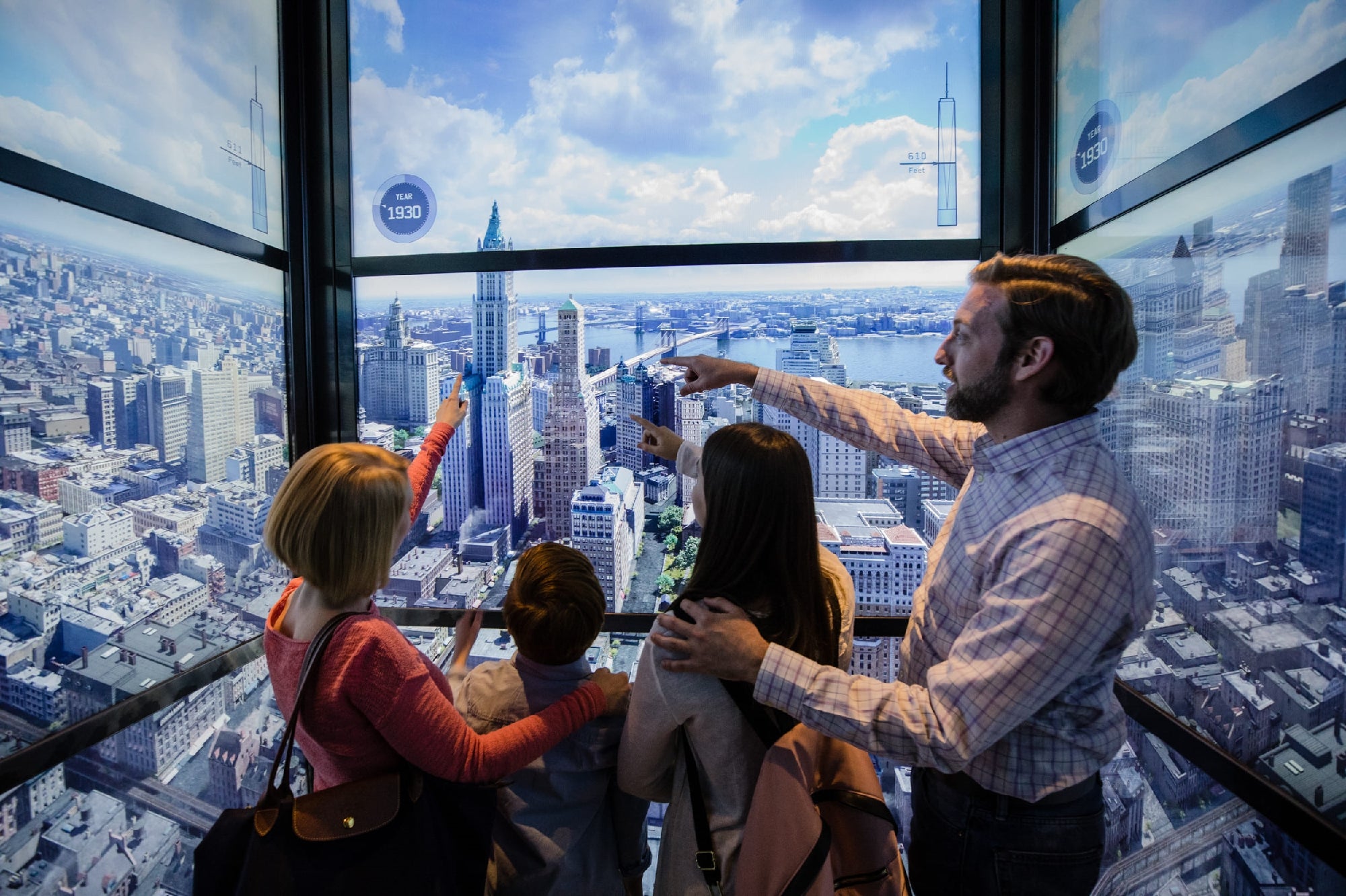 A family of four stands in an observation area with panoramic cityscape views, pointing and looking out at the skyline through floor-to-ceiling windows.