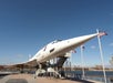 A retired Concorde supersonic jet is displayed on an outdoor platform beside a river, with stairs for visitors and American flags nearby.