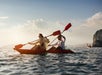 Two people paddle a red kayak on the open water at sunset, with distant cliffs and a boat visible in the background.