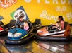 Two adults and two children ride bumper cars indoors, with the cars about to collide near a yellow wall labeled “Bumper Car.”.