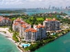 Aerial view of waterfront condominiums with red roofs on an island, surrounded by water, with a city skyline visible in the distance.